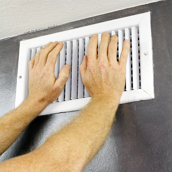 Two adult hands placed over an out air vent register of a central heating system on a gray wall near a white ceiling.